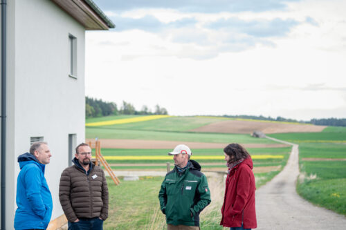 Vier Personen stehen vor einem modernen Gebäude am Rand eines Feldwegs und diskutieren.