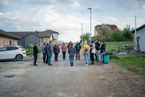 Eine Gruppe von etwa 15 Personen steht in einem Kreis auf einer unbefestigten Straße in einem Wohngebiet.