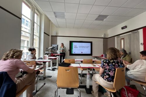 Eine kleine Gruppe an Menschen sitzt in einem Klassenzimmer. Eine Frau geht vor einem Fernseher und Präsentiert.