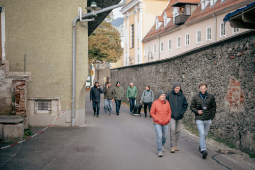 Eine Gruppe, bestehend aus 8 Personen, geht eine Gasse entlang einer alten Stadtmauer entlang.