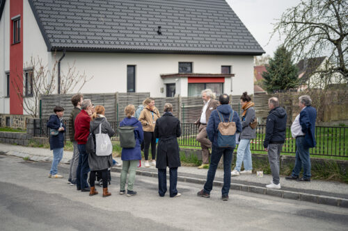 Eine Menschengruppe vor einem Einfamilienhaus auf einer Straße stehend.