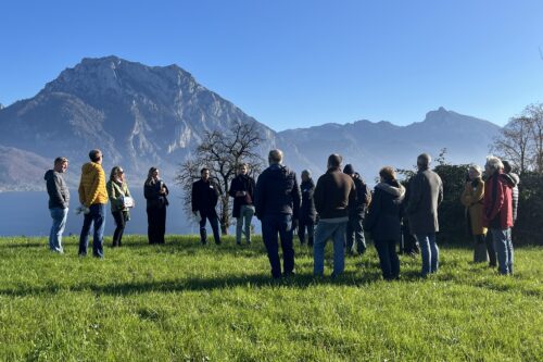 Eine Menschengruppe steht auf einer Feldwiese. Im Hintergrund ein Bergpanorama